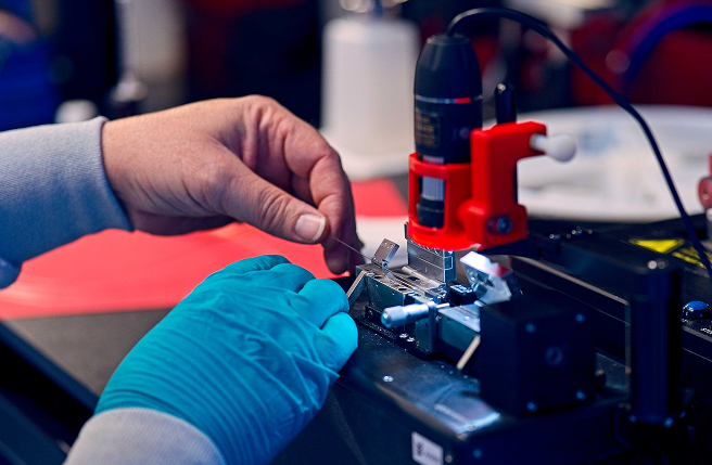 Deux mains effectuant un travail de précision sur un petit dispositif électronique, à l’aide d’un outillage spécialisé et sous un éclairage directionnel.