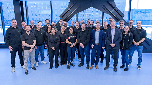 LumIR Lasers team gathered in a bright indoor space, posing in front of a lattice shaped metal structure, with large windows looking out onto buildings in the background.