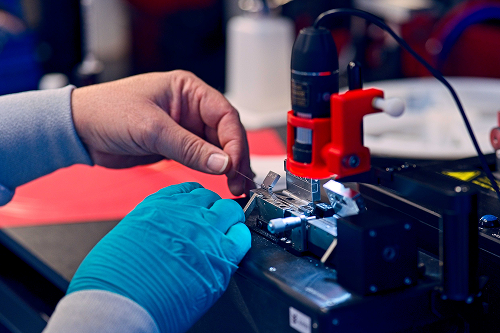 Two hands performing precision work on a small electronic device, using specialized tools and working under directional lighting.