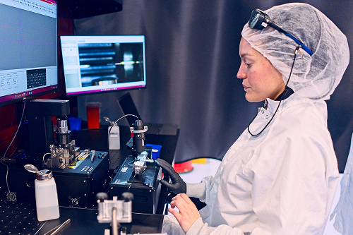 Person in laboratory attire operating precision equipment in front of monitors displaying technical data.