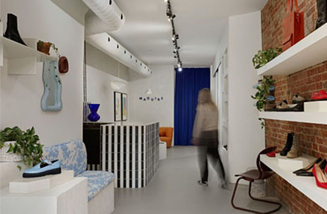 Intérieur d’une boutique avec étagères présentant des chaussures et sacs, un comptoir et une personne marchant vers le fond.