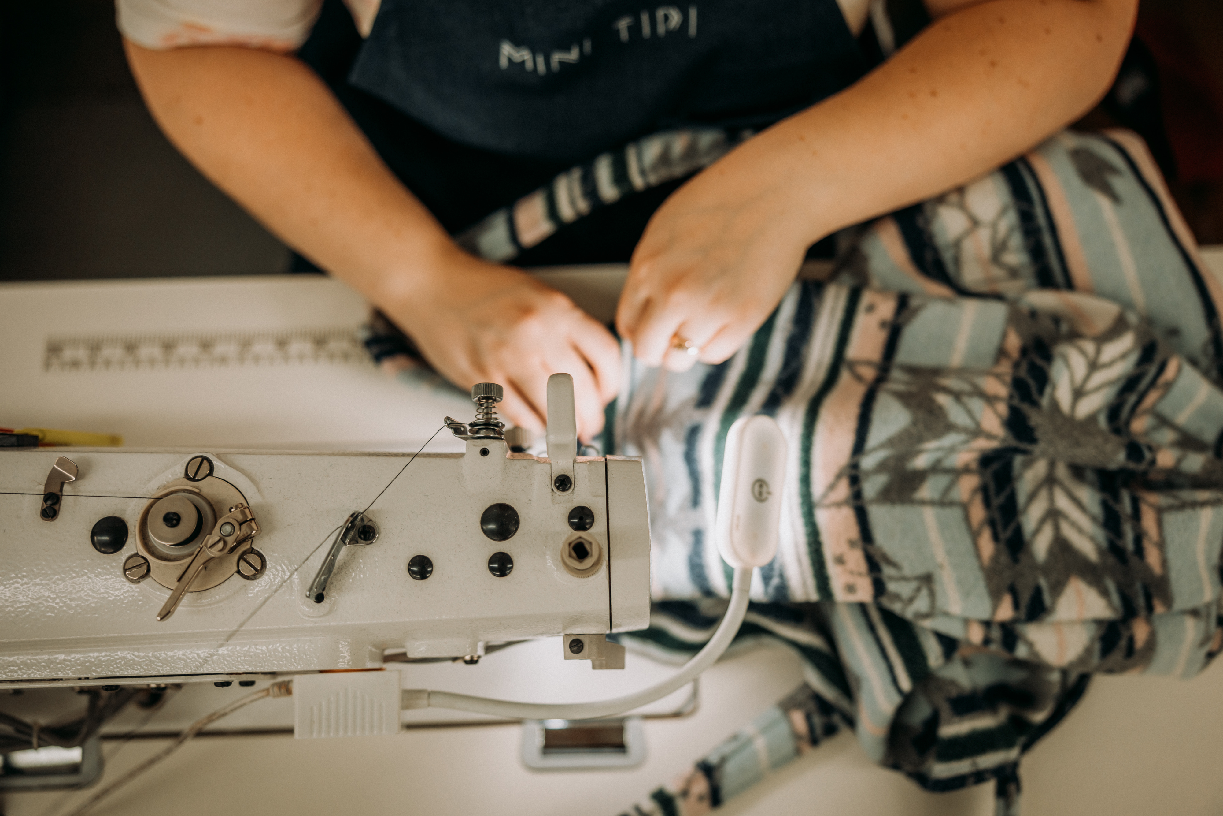 A close-up shows the hands of a worker seated at a sewing machine, finishing a MINI TIPI product decorated with traditional indigenous patterns.