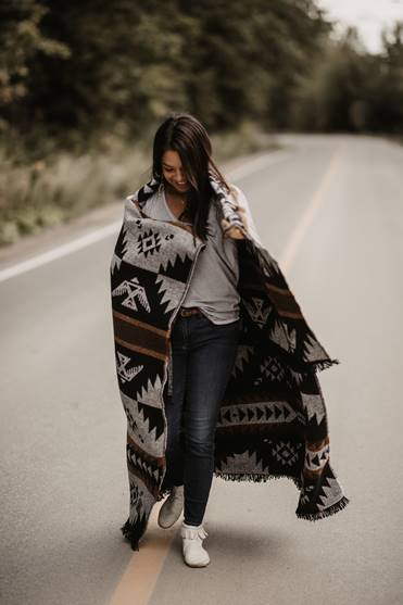 A woman on a deserted road is wrapped in a blanket decorated with traditional native motifs.