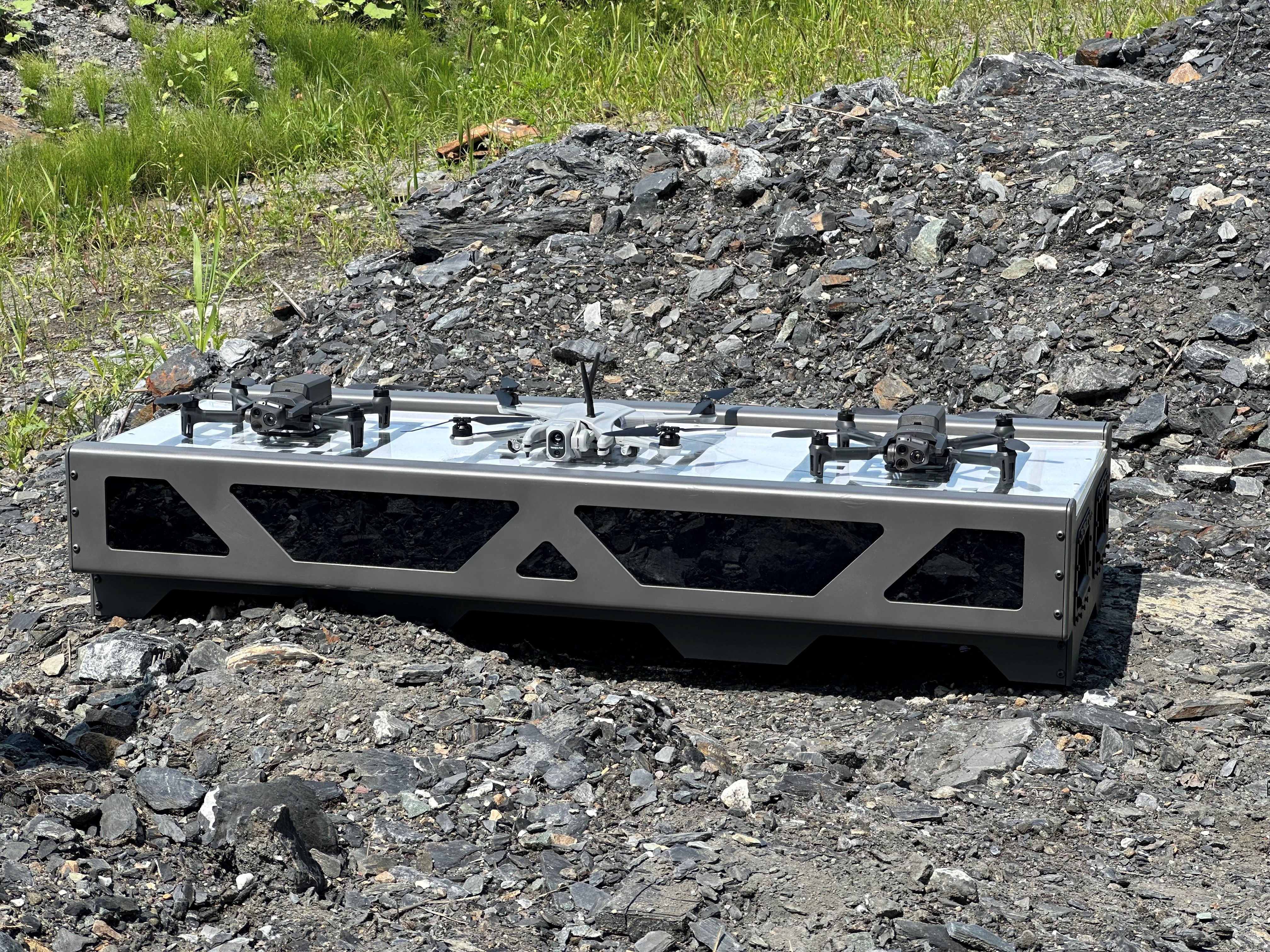 Trois drones sur une station de recharge autonome déposée sur un terrain rocailleux. 