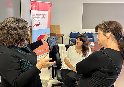 Three women chat animatedly around a banner that reads in french “Funding for impact businesses - Evol”.