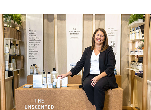 Anie Rouleau, vêtue d’un complet noir et d’un t-shirt blanc avec la mention The Unscented Company, est assise sur un bloc de liège avec les produits de la gamme The Unscented Company à ses côtés.