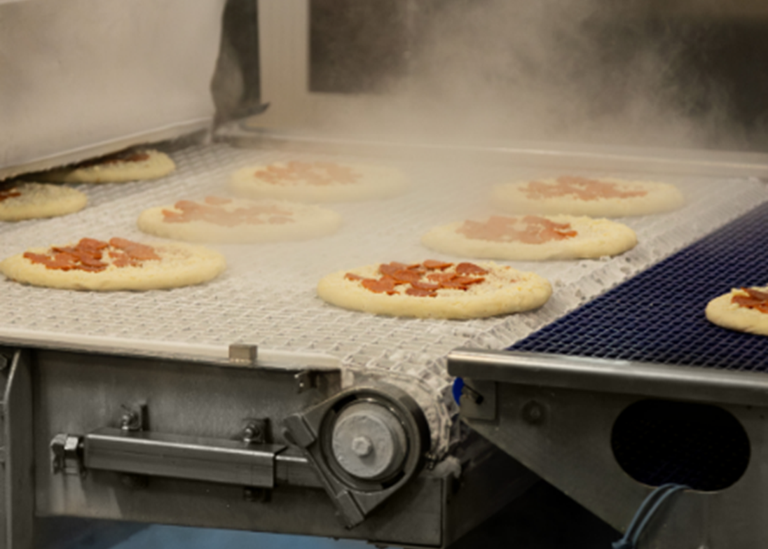 Chaîne de production alimentaire industrielle où des pizzas garnies sont transportées sur un convoyeur métallique, avec dégagement de vapeur dans un environnement automatisé.