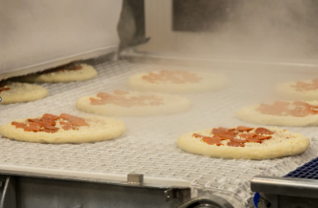 Chaîne de production alimentaire industrielle où des pizzas garnies sont transportées sur un convoyeur métallique, avec dégagement de vapeur dans un environnement automatisé.