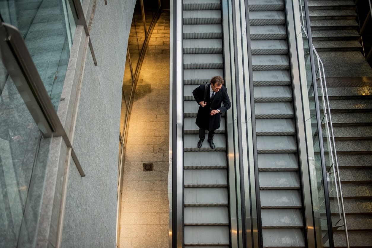 A businessman descends an escalator, looking at his watch. The building looks empty. The photo is taken from above, and other staircases are visible to the right of the man, accentuating the impression of solitude.