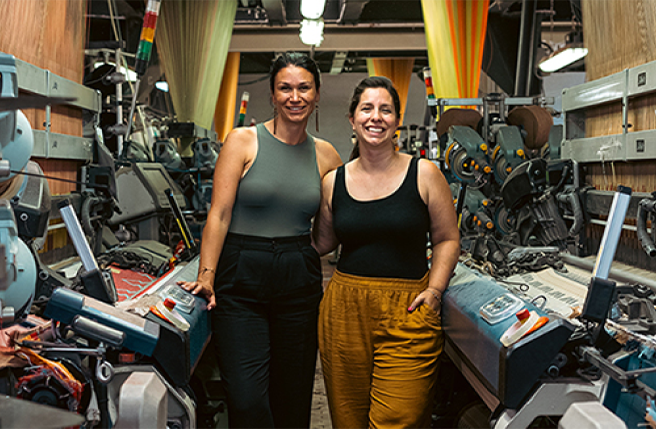 Mélanie Bernard et Trisha Pitura posent au milieu de l’atelier de production de MINI TIPI, avec des métiers à tisser industriels de part et d’autres de la photo.