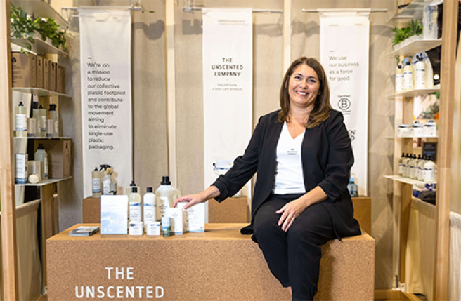 Anie Rouleau, vêtue d’un complet noir et d’un t-shirt blanc avec la mention The Unscented Company, est assise sur un bloc de liège avec les produits de la gamme The Unscented Company à ses côtés.