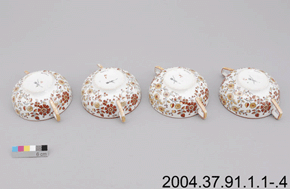 Colour photo of four patterned tea-cups with doubled handles, showing trademark on each base with a colour scale and 2004.37.91.1.1-.4