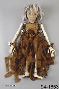 Close-up of a doll with skeletal arms/legs in a loose brown dress with the number 94-1653