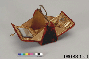 Colour photo of the interior of a red and gold handbag, with a padded tan interior and small mirror with a colour scale and the text 980.43.1 a-f