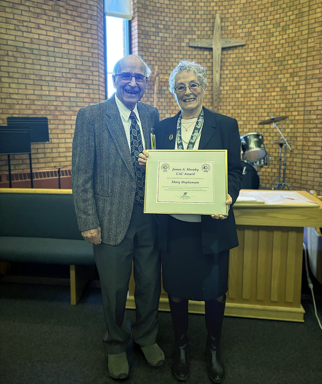 Deux personnes se tiennent à l'intérieur d'une église avec des murs en briques et une grande croix en bois sur le mur derrière elles. Elles tiennent un certificat encadré qui indique James A. Murphy CAC Award présenté à Mary Stephenson.