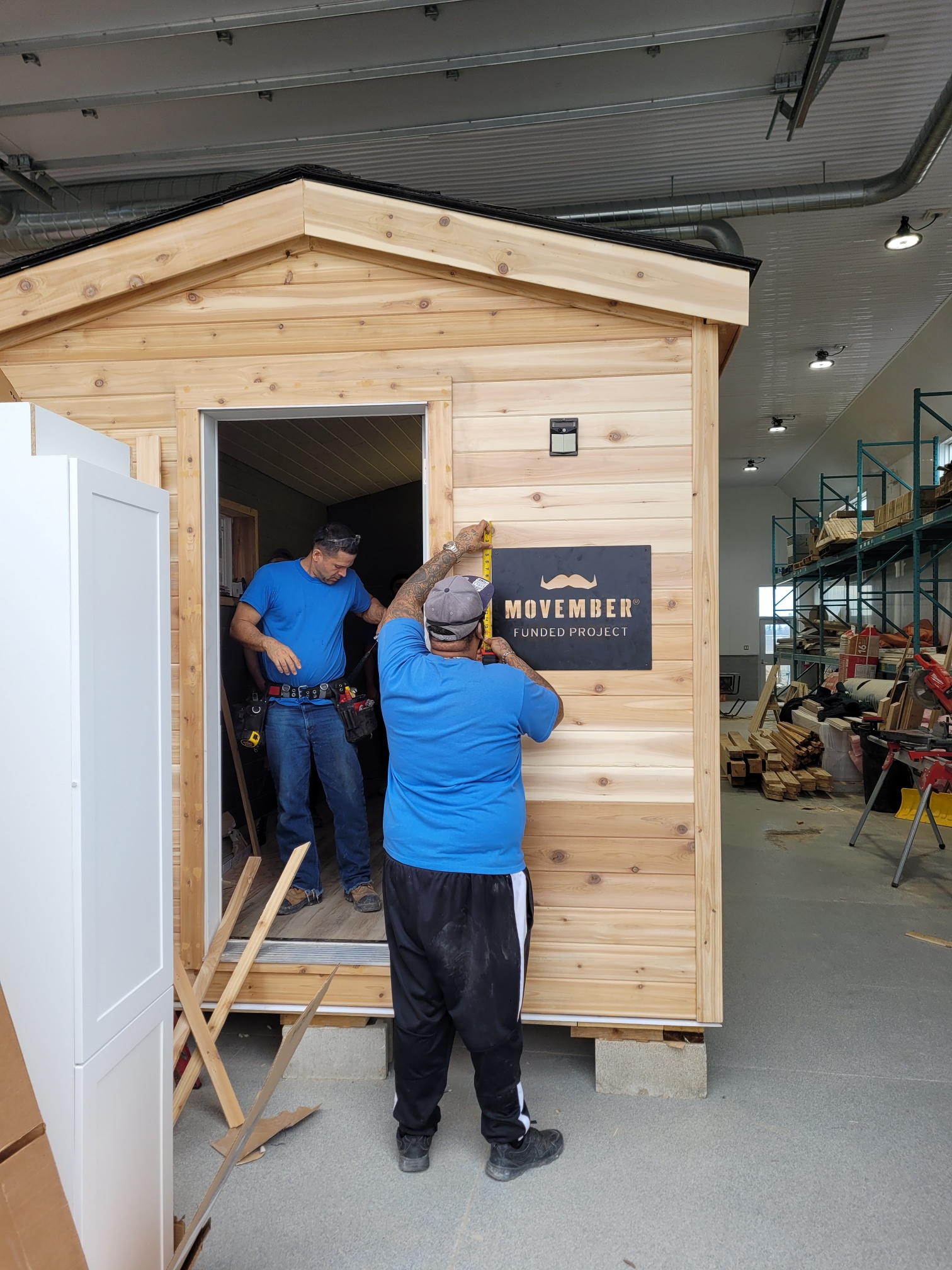 Deux hommes travaillent sur une minimaison dans l’entrepôt de Construction CORCAN à l’Établissement de Collins Bay. L’un d’eux se tient dans le cadre de porte et l’autre mesure une affiche sur laquelle on peut lire : Movember Funded Project (Projet financé par Movember).