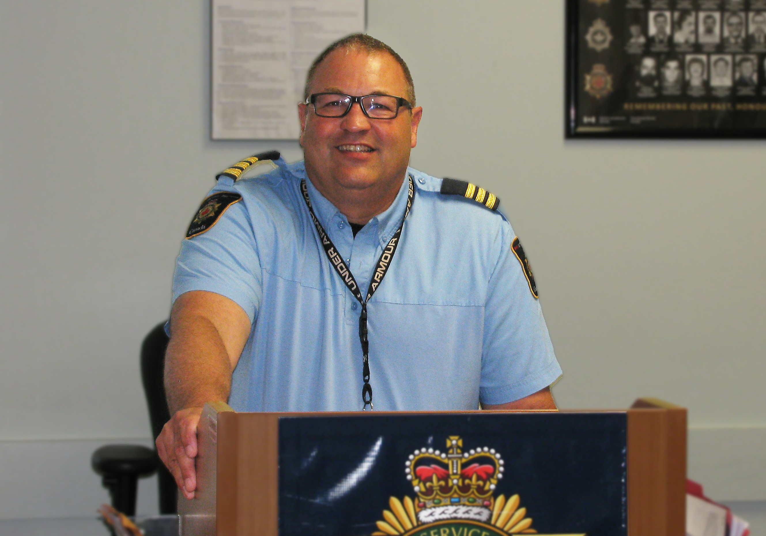 Troy Talbot souriant en uniforme derrière un podium. En arrière est l'Énoncé des valeurs du Service correctionnel du Canada (SCC) et un mémorial au personnel passé du SCC.