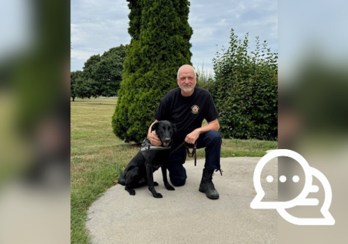 Darrell Scott kneeling next to his Detector Dog Jaxx, a black Labrador, on a leash