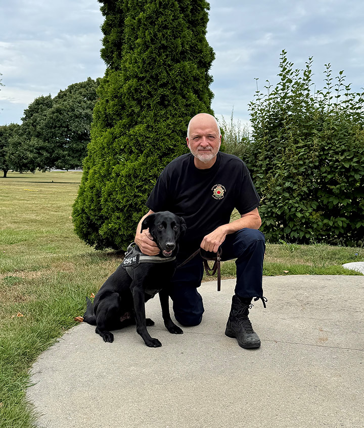 Darrell Scott à genoux à côté de son chien détecteur Jaxx, un Labrador noir, en laisse
