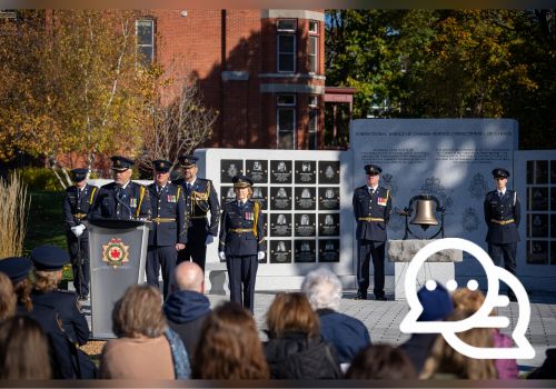 Les dirigeants et le personnel du SCC sont placés sur un podium et devant un grand mur commémoratif en granit avec des plaques et une cloche devant.