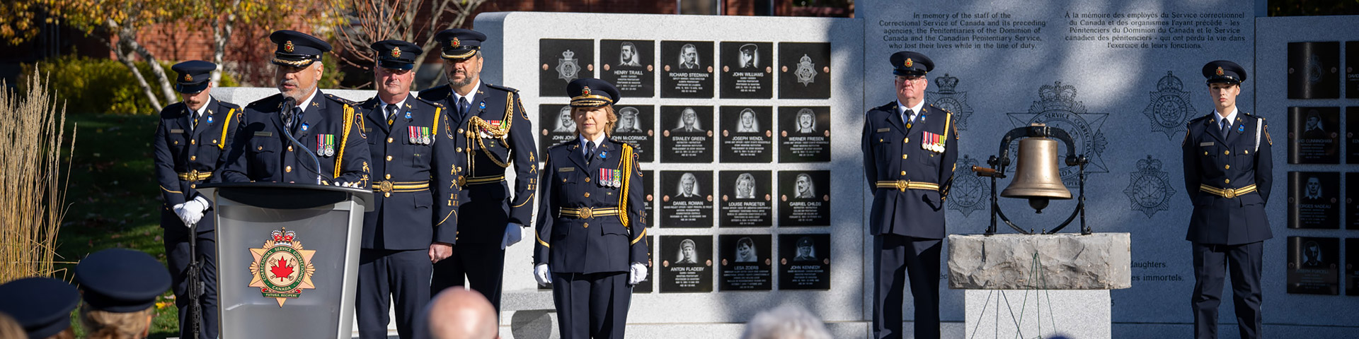 CSC executives and staff at a podium and in front of a large granite memorial wall with plaques and a bell in front of it.