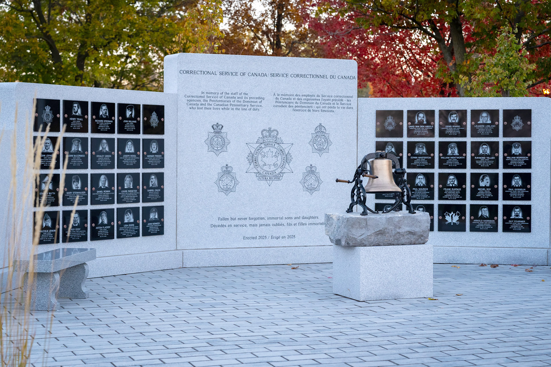 A large granite wall memorial with plaques in front of a bell and stone benches.