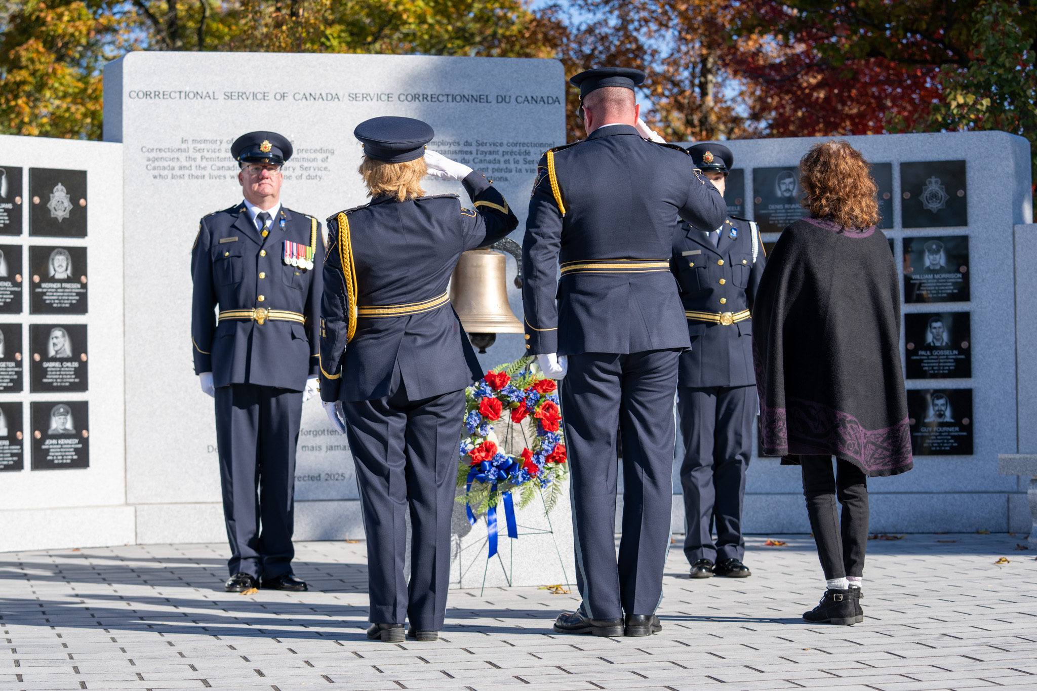 A woman and a man saluting a wreath in front of a large granite wall memorial and bell in CSC uniforms beside a woman watching on. Two other people in CSC uniforms stand on either side of the bell.
