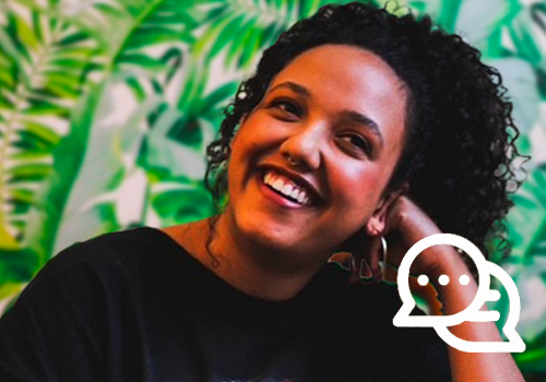 A young woman with curly black hair in a high ponytail and a black t-shirt laughs off to the side. She is resting her face on her hand at a white table, in front of leafy green wallpaper.