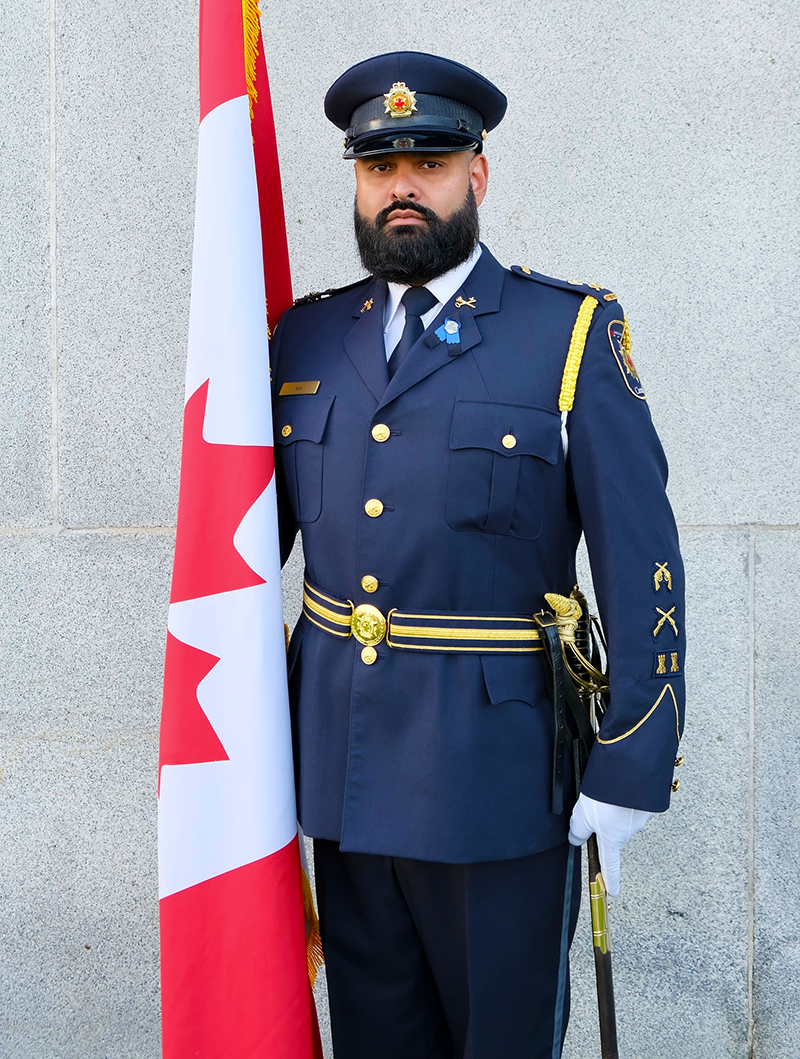 Un gestionnaire en uniforme du Service correctionnel Canada, Chanveer Rai, porte le drapeau canadien. Il porte l’uniforme de cérémonie de la garde d’honneur bleu foncé avec des accents dorés, un chapeau, des gants blancs et une épée de cérémonie.
