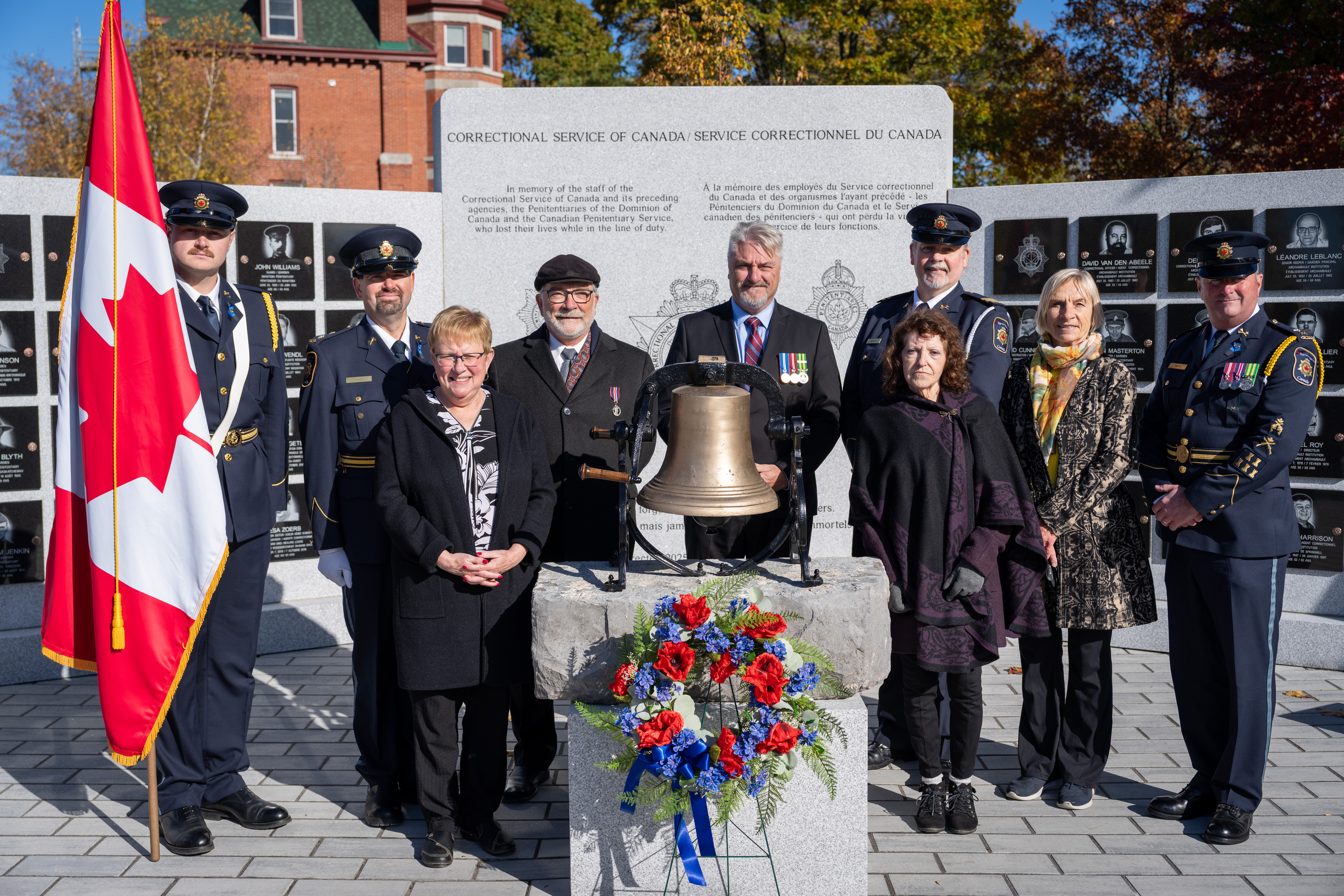The eight committee members standing around the bell and wreath in front of the memorial. Jacob Ritchie, son of committee member Scott Ritchie, stands on the left holding a Canadian flag.