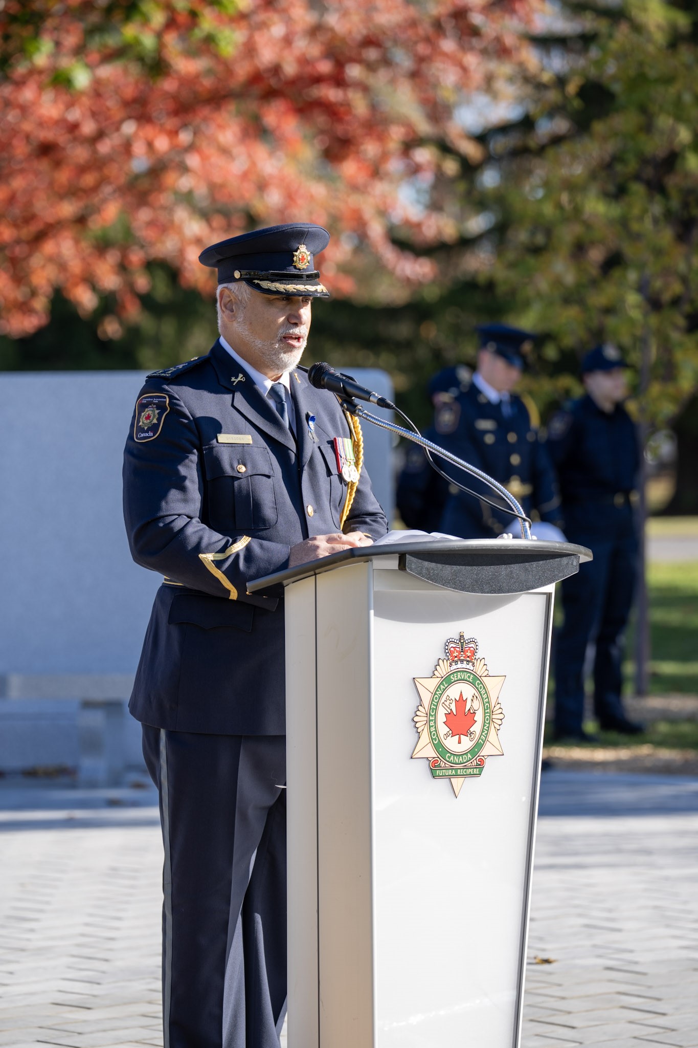 RDC Kevin Snedden in uniform speaks outdoors in front of the memorial at a podium adorned with the CSC badge.