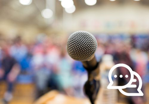 A close-up image of a microphone on a podium with a blurred audience in the background.
