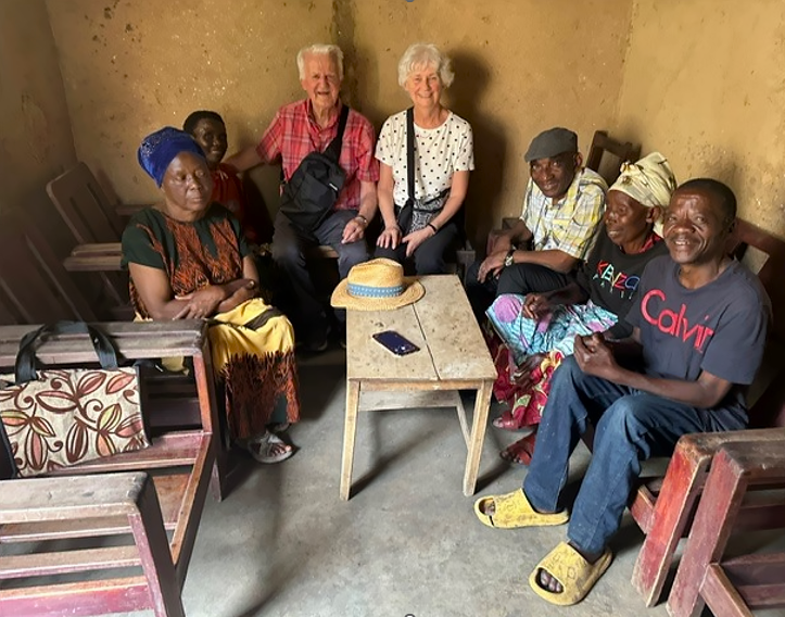 Seven people sit together around a small wooden table inside a simple room with earthen walls, smiling as they gather for a group meeting.