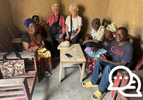Seven people sit together around a small wooden table inside a simple room with earthen walls, smiling as they gather for a group meeting.