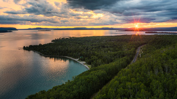 Vue aérienne d'un coucher de soleil sur une baie et le rivage boisé ; une voie ferrée traverse la forêt. Le ciel est d'ailleurs assez nuageux. .