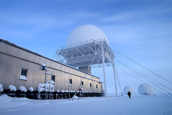A large white geodesic dome sits on a platform above a rectangular building with metallic siding. Two smaller white geodesic domes are in the background. A person in silhouette walks in the distance. The foreground is a snow-covered field.