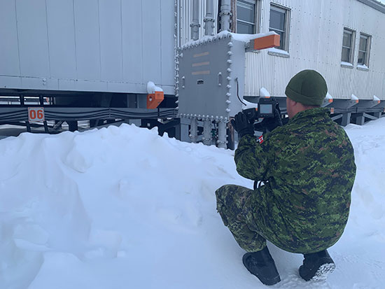 A Canadian Armed Forces member points a thermal camera at the outside of a building with a pile of snow in front of it.