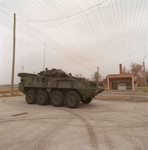 A large, green light armoured vehicle is parked underneath a series of parallel wires suspended by poles in an outdoor facility.