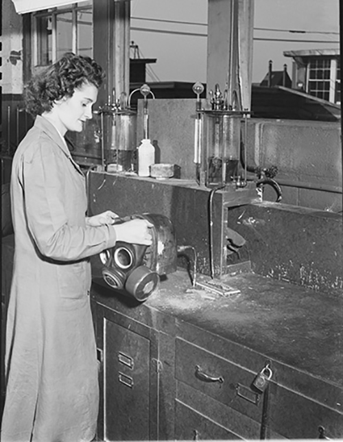A black and white photo of a woman in a lab coat handling a gas mask in a laboratory.