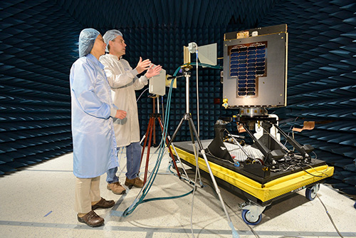 Deux scientifiques discutent d'un satellite monté sur une table à roulettes à l'intérieur d'une salle insonorisée.