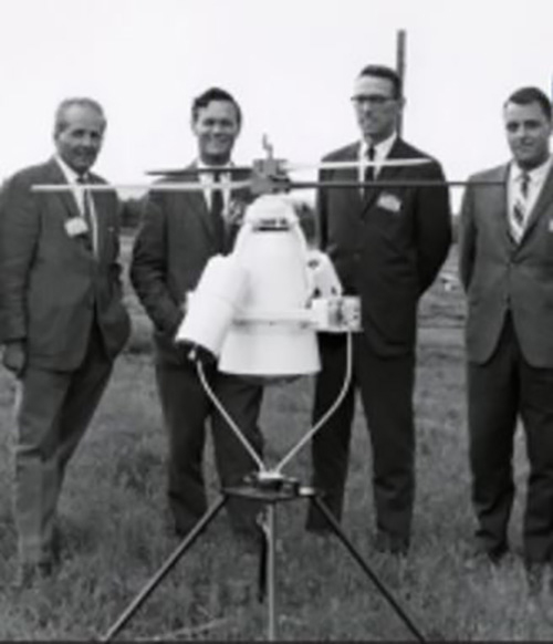 Photo en noir et blanc montant quatre hommes en habit posant derrière un véhicule aérien sans pilote dans un champ.