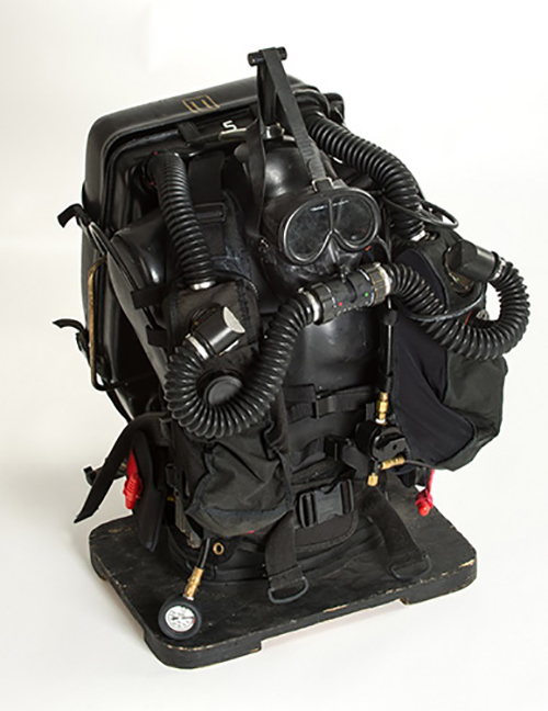 Photo d'un dispositif respiratoire submersible entièrement noir doté de tubes respiratoires connectés au masque et d'un large étui au dos posé sur une planche noire contre un fond blanc.