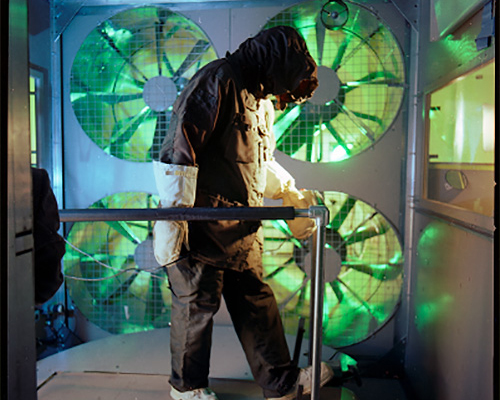 A person covered from head to toe in military cold weather clothing walks on a treadmill in front of four large fans, with green light shining through the blades.