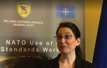 A woman in front of a sign with the NATO symbol and text “Hellenic National Defence General Staff. NATO Use of Civil Standards workshop.”