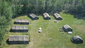 Des tentes militaires vertes sont disposées en demi-cercle dans un champ entouré de forêt.