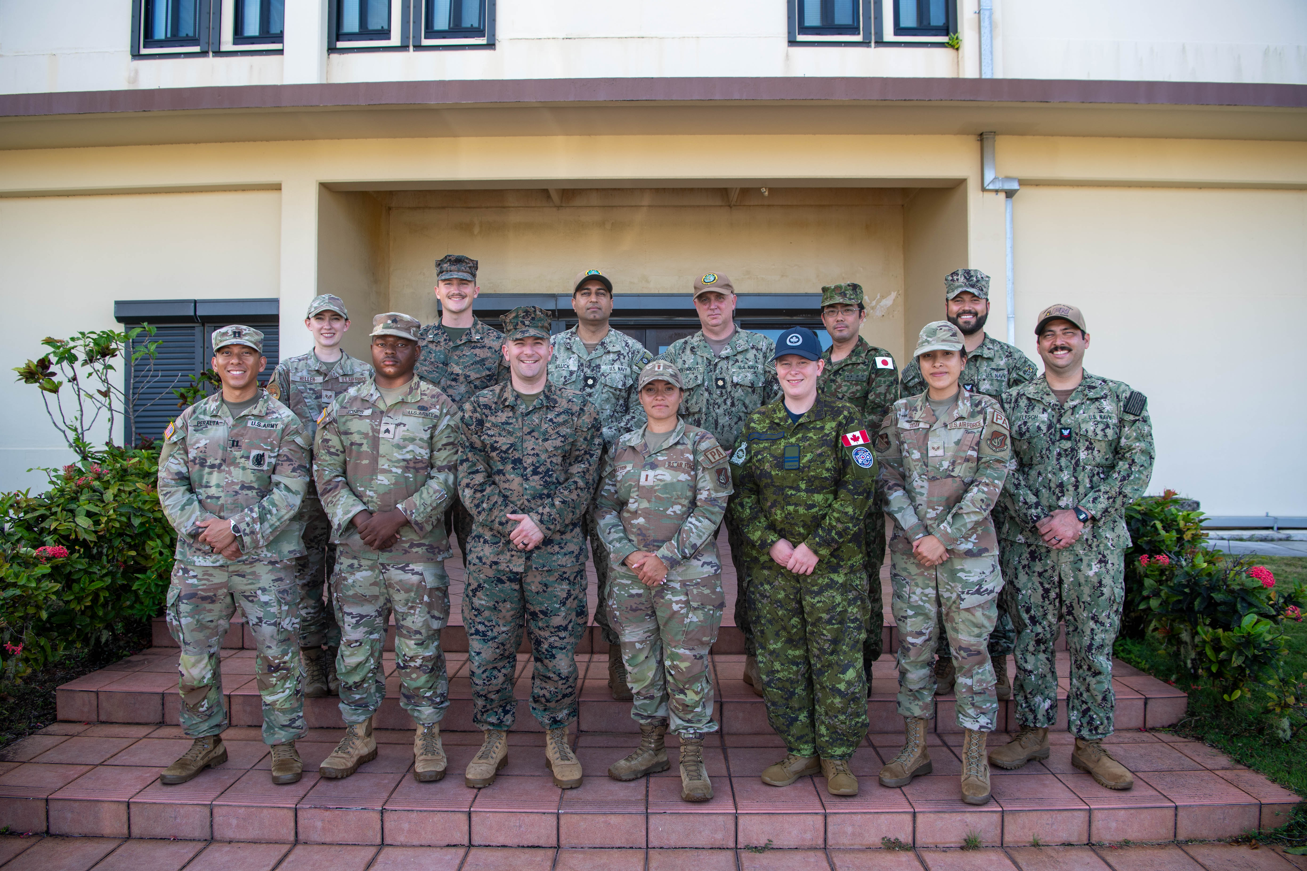 Thirteen uniformed members of the Canadian, Japanese, and United States armed forces stand together on the front steps of a building in Guam.