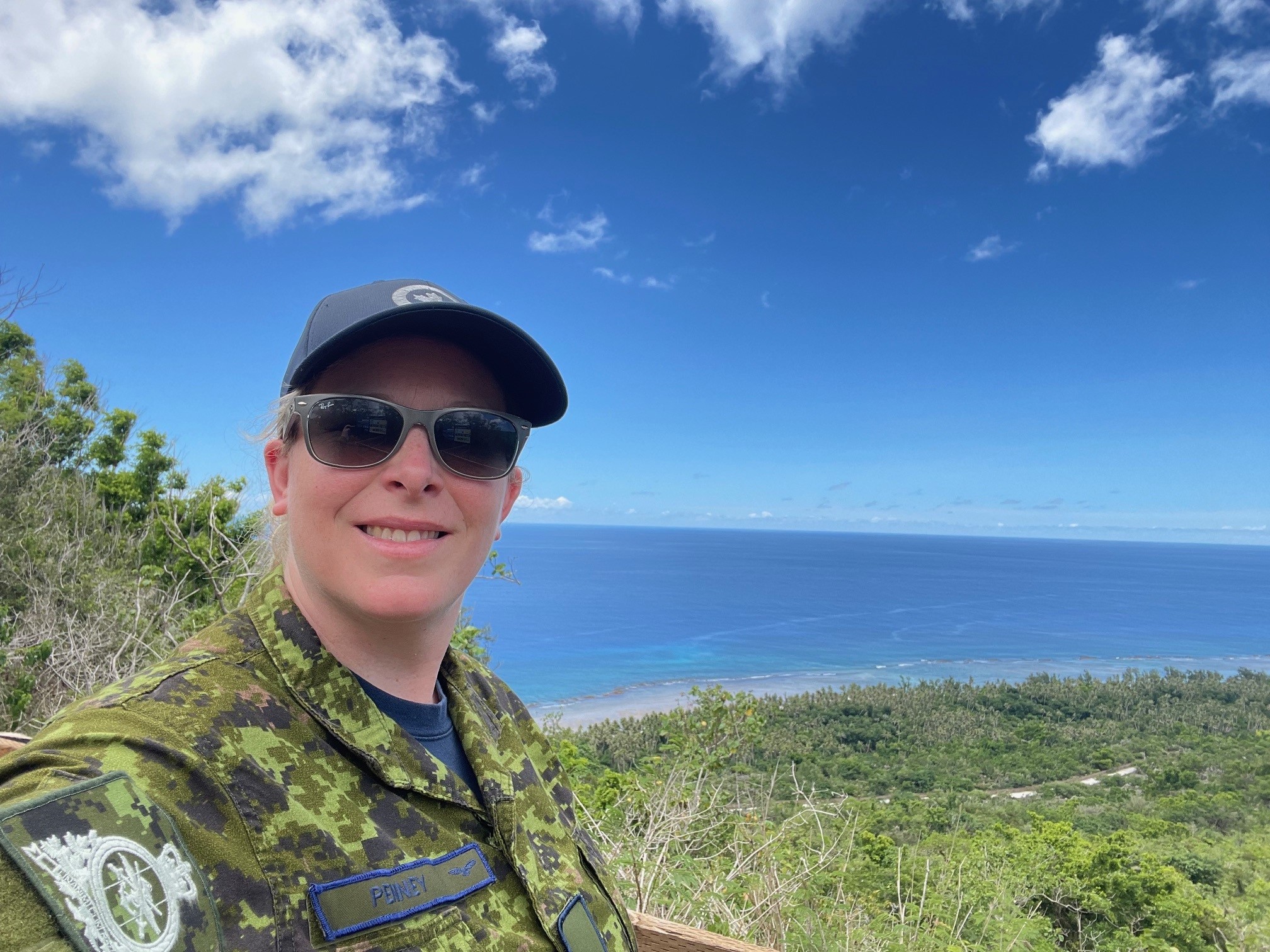 Captain Kylie Penney poses for a selfie in uniform in Guam with green trees, blue ocean, and clear sky in the background.