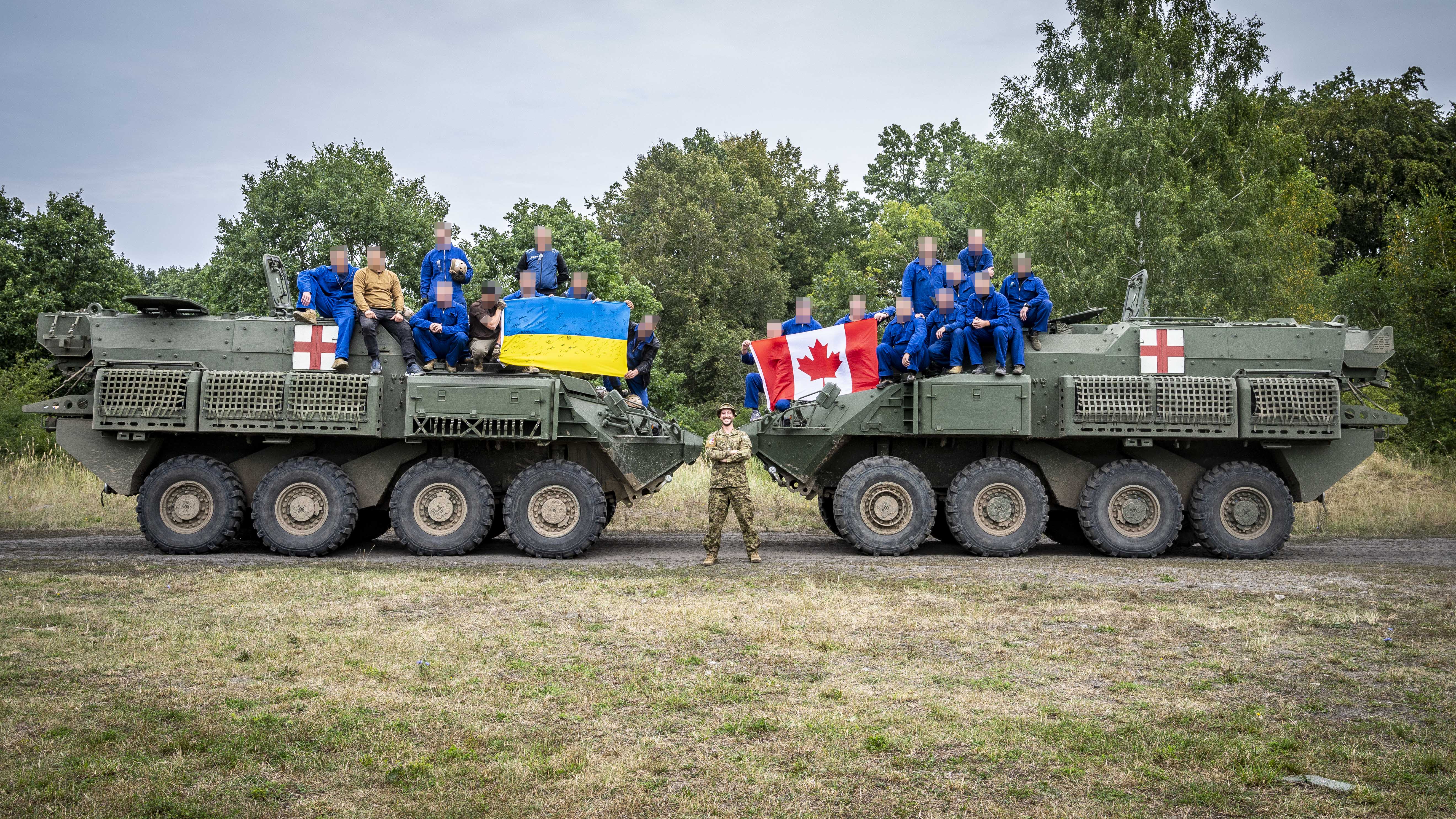 Two groups of Armed Forces of Ukraine members sit on top of two separate tanks. One group is holding a Ukrainian flag while the other holds a Canadian flag. The two tanks are facing each other in a field with a forest behind them.