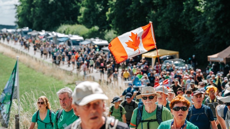 A large group of people walks along a road beside a grassy field during an outdoor event. Many wear hats and backpacks. A prominent Canadian flag is held up in the center. Trees, tents, and vehicles line the roadside in the background.