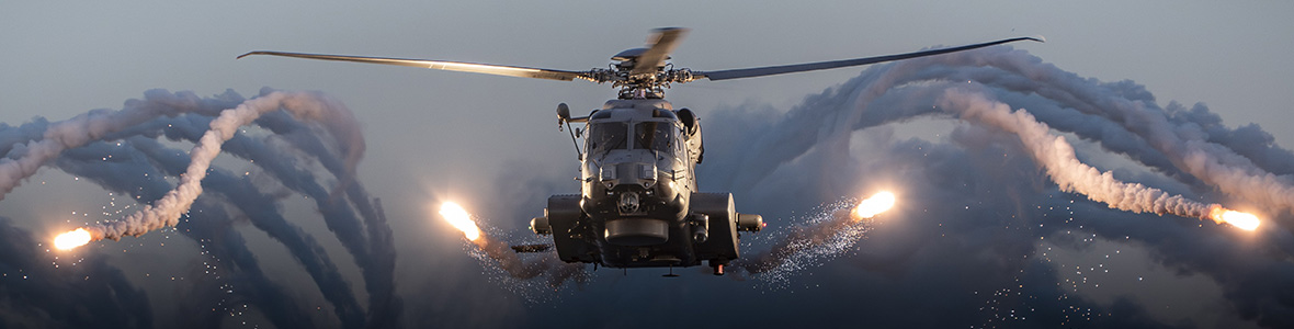 A military helicopter releases symmetrical flares while flying through a cloudy sky, creating curved trails of smoke and light
